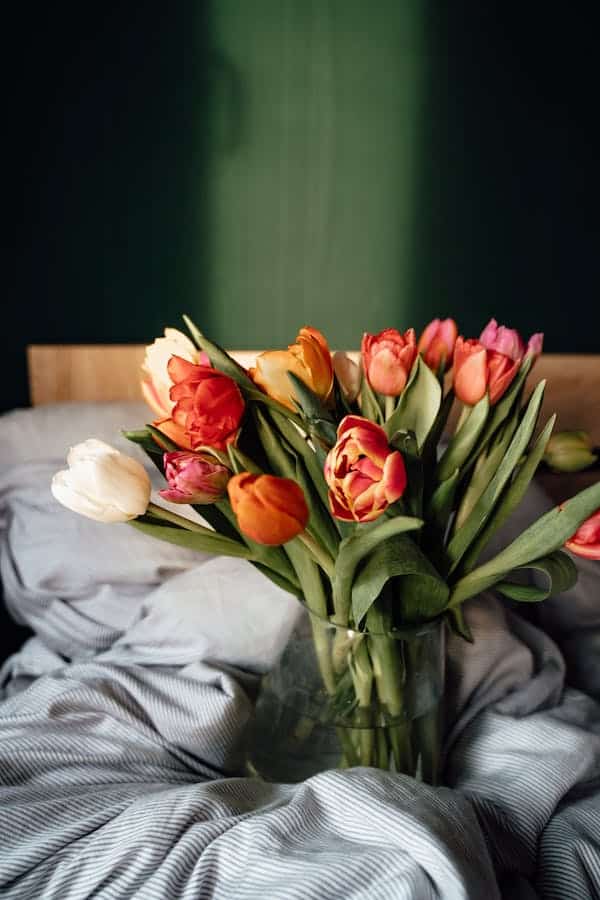 Bouquet de tulipes colorées dans un vase en verre posé sur un lit avec des draps rayés gris. Ambiance cosy.