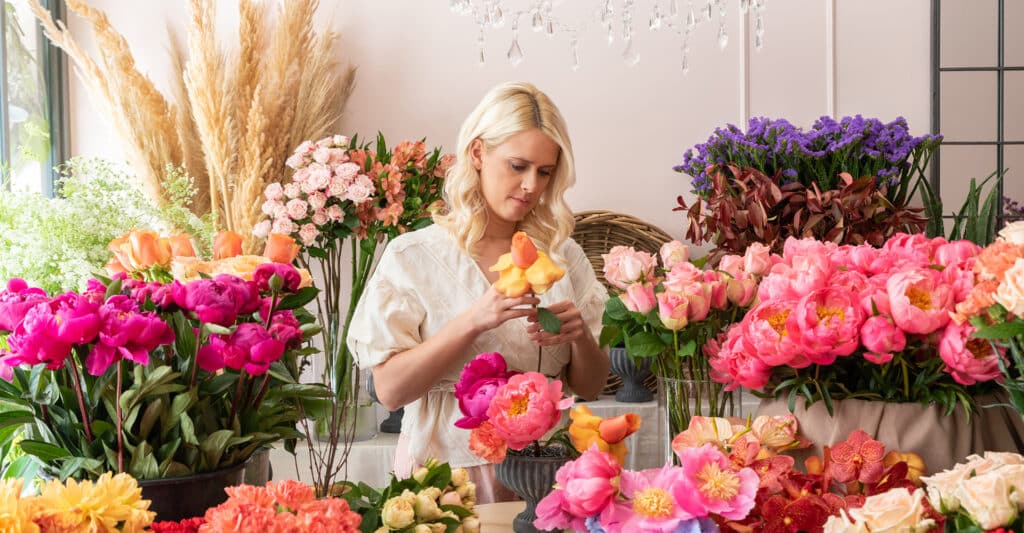 Blonde femme arrangeant des fleurs dans un atelier. Peonies roses, roses peche, statice violet, herbe de la pampa.