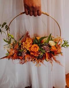 Main tenant un bouquet cerceau de mariage avec roses orange, feuillage automnal et plumes sur fond blanc.