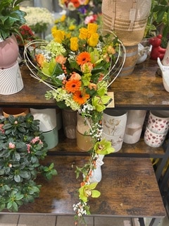 Arrangement floral en cœur avec roses jaunes, gerberas orange et orchidées vertes retombantes sur étagère en bois.