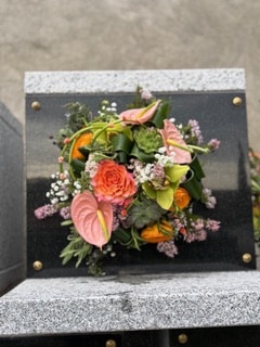 Bouquet funéraire coloré avec rose orange, anthuriums roses et orchidées sur une plaque de granit noir.