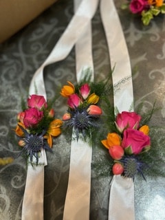 Trois corsages de poignet avec roses roses, chardons bleus et fleurs orange sur des rubans en satin blanc.