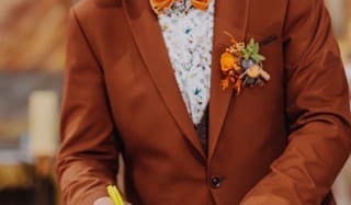 Homme en costume terracotta, nœud papillon orange et boutonnière automnale signant un document de mariage.