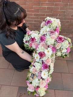 Femme tenant une grande croix florale composée de roses et de gerberas roses, blancs et violets.