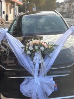 Voiture de mariage noire décorée de tulle blanc et d'un bouquet de roses roses et blanches sur le capot.
